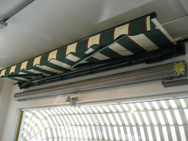 Markise mit grün-beigem Streifenmuster als Sonnenschutz über einem Fenster im Innenraum.