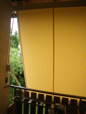 Gelbe Markise mit Streifen spendet Schatten auf einem Balkon mit Holzgitter und Ausblick ins Grüne.